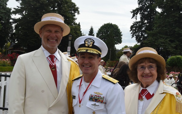 Three individuals pose together at what appears to be a formal outdoor ceremony, likely a Rose Festival or similar civic event. Two individuals wear matching cream/white suits with straw boater hats labeled 'ROYAL' and gold ceremonial capes, while the central figure wears a U.S. Navy dress white uniform with multiple ribbons and medals. The naval officer holds a framed certificate or proclamation document.