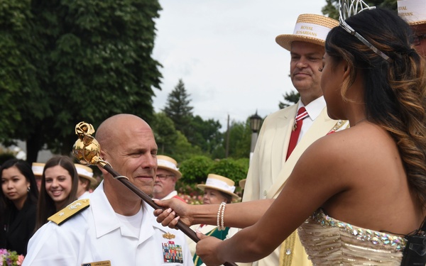 A U.S. Navy admiral in white dress uniform kneels while a young woman in a formal gold/silver gown touches his shoulder with a ceremonial scepter topped with a golden rose. The setting appears to be an outdoor ceremony associated with a Rose Festival, evidenced by the 'Royal Rosarians' insignia visible on the jackets of attendants in the background. Spectators seated in chairs observe the formal knighting or coronation-style ceremony.