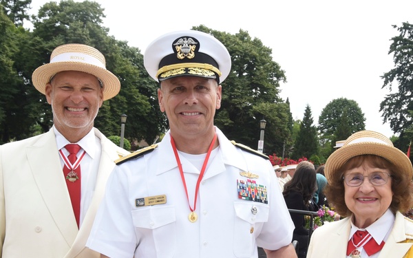 Three individuals pose together at what appears to be an outdoor ceremonial event. A U.S. Navy officer in white dress uniform holds a framed certificate, flanked by two people wearing cream/white suits with straw boater hats and yellow ceremonial sashes. The setting appears to be a formal garden or park with a water feature visible in the background.