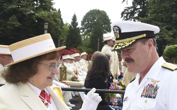 A formal outdoor ceremony is taking place, likely the Portland Rose Festival based on the visible 'ROS' text on the woman's hat band. A woman in a cream blazer, straw boater hat, and white gloves is presenting a framed document or certificate to a U.S. Navy officer in dress whites. The background shows rows of attendees in matching cream and white attire, with rose gardens and trees visible.