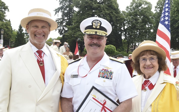 Three individuals pose together at what appears to be a formal outdoor ceremony or parade. A U.S. Navy officer in dress whites holds a framed certificate tied with a red ribbon, flanked by two civilians wearing cream-colored suits and straw boater hats with red accessories. An American flag is visible in the background along with a crowd of similarly dressed attendees.