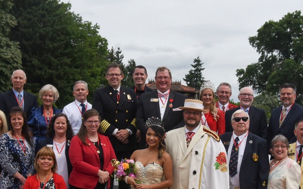 A large formal group photograph featuring approximately 35 individuals posed together outdoors in what appears to be a ceremonial or celebratory event. At the center are two prominent figures dressed in royal/ceremonial attire — a young woman in a gold gown wearing a crown and a man in a white suit with a decorative hat, suggesting a festival queen and king or similar honorary roles. The group includes military personnel in white naval uniforms, individuals wearing red sashes and medals, and civilians in formal attire, set against a backdrop of green trees and a garden setting.