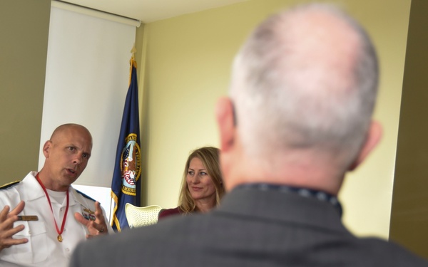 A U.S. Navy officer in white dress uniform is speaking and gesturing with his hands during what appears to be a meeting or briefing. He is seated across from at least two other individuals, one of whom is visible from behind in the foreground. A blue naval flag is visible in the background.