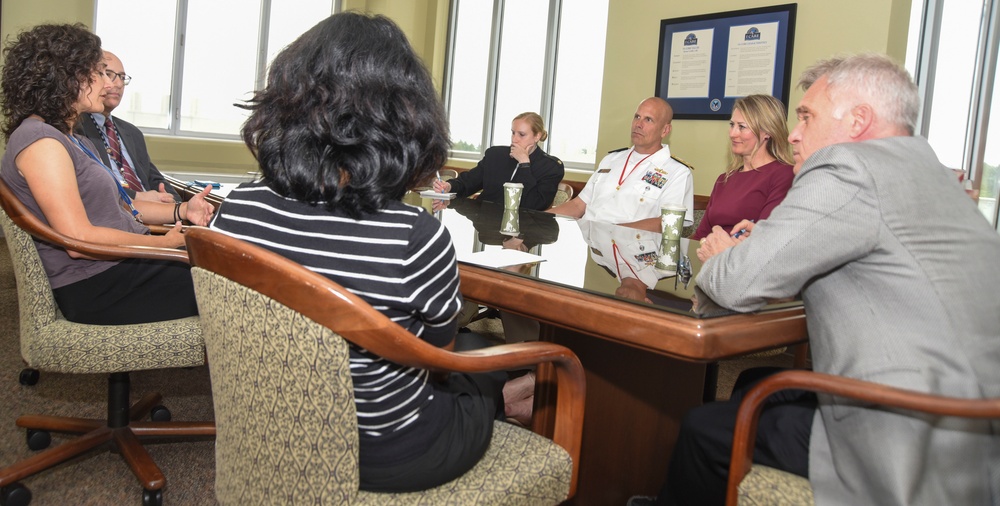 A formal meeting is taking place around a large conference table in what appears to be a government or military office setting. Seven individuals are seated around the table, including one person in a white naval uniform with medals and insignia. Framed documents or certificates are visible on the wall in the background.