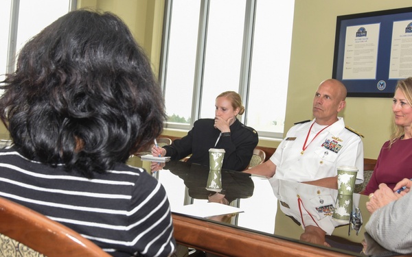 A formal meeting is taking place around a large conference table in what appears to be a government or military office setting. Seven individuals are seated around the table, including one person in a white naval uniform with medals and insignia. Framed documents or certificates are visible on the wall in the background.