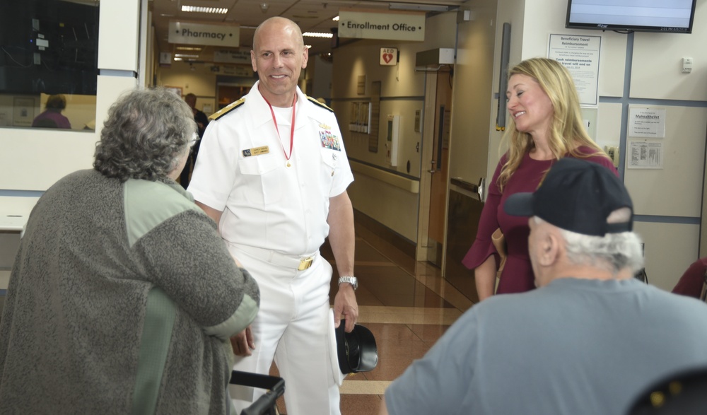 A U.S. Navy officer in white dress uniform is greeting elderly visitors or patients in what appears to be a military medical facility or VA hospital. He is accompanied by a woman in a burgundy dress, and they appear to be engaging with an older couple. Signs in the background indicate a Pharmacy and Enrollment Office.