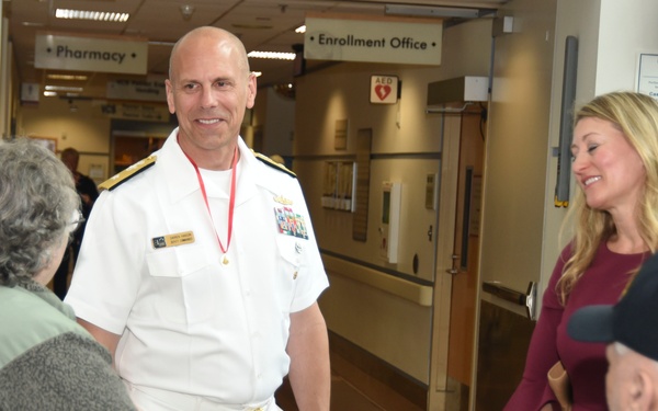 A U.S. Navy officer in white dress uniform is greeting elderly visitors or patients in what appears to be a military medical facility or VA hospital. He is accompanied by a woman in a burgundy dress, and they appear to be engaging with an older couple. Signs in the background indicate a Pharmacy and Enrollment Office.