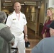 A U.S. Navy officer in white dress uniform is greeting elderly visitors or patients in what appears to be a military medical facility or VA hospital. He is accompanied by a woman in a burgundy dress, and they appear to be engaging with an older couple. Signs in the background indicate a Pharmacy and Enrollment Office.