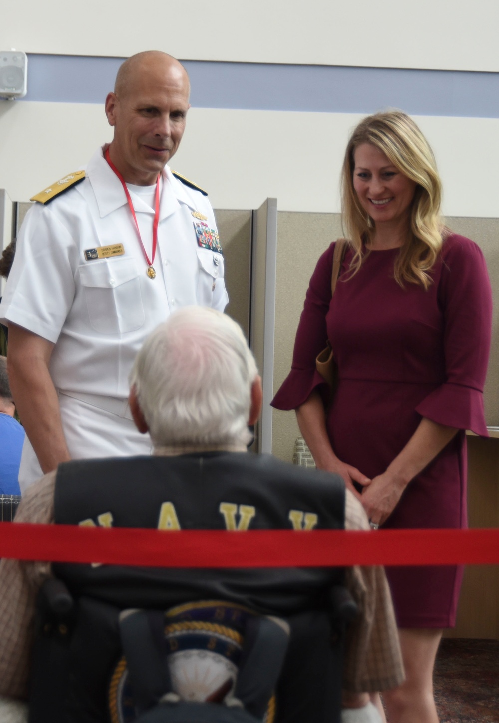 A U.S. Navy officer in white dress uniform wearing a red-ribboned medal is standing alongside a woman in a burgundy dress, both smiling and engaging with an elderly veteran seated in a wheelchair. The veteran is wearing a black jacket and cap with 'NAVY' lettering. The setting appears to be an indoor facility, possibly a veterans' event or ceremony.