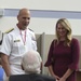 A U.S. Navy officer in white dress uniform wearing a red-ribboned medal is standing alongside a woman in a burgundy dress, both smiling and engaging with an elderly veteran seated in a wheelchair. The veteran is wearing a black jacket and cap with 'NAVY' lettering. The setting appears to be an indoor facility, possibly a veterans' event or ceremony.