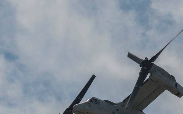 A military tiltrotor aircraft, identifiable as a Bell Boeing V-22 Osprey, is photographed from below against a partly cloudy sky. The aircraft bears the markings '04' on its fuselage and 'EM 04' on its tail, indicating U.S. Marine Corps affiliation. The rotors are in a transitional or forward-flight configuration, angled upward from the wingtips.