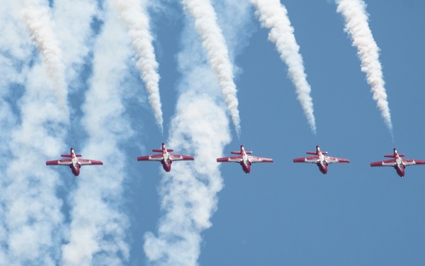 Six red and white jet aircraft fly in a diagonal line formation against a clear blue sky, each trailing white smoke streams. The aircraft appear to be part of an aerobatic display team performing a synchronized formation flight. The smoke trails create a dramatic parallel pattern descending from upper left to lower right.