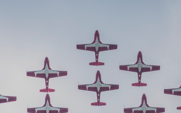 A formation of nine red and white military jet aircraft are performing an aerobatic display, viewed from below against an overcast grey sky. The aircraft are arranged in a diamond/arrow formation typical of precision aerobatic teams. Several of the aircraft are emitting white smoke trails, indicating an active airshow performance.