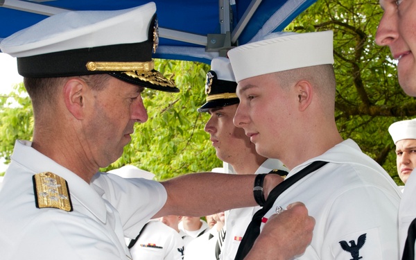 A senior U.S. Navy officer, identifiable by gold shoulder boards and a combination cover with gold oak leaf scrambled eggs, is pinning a medal or award onto an enlisted sailor wearing white dress uniform. The sailor receiving the award displays a Petty Officer First Class rating badge on his left sleeve. The ceremony appears to be taking place outdoors under a blue canopy tent with additional Navy personnel standing in formation in the background.