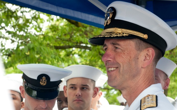 A U.S. Navy officer in dress white uniform with gold insignia and a scrambled-egg cap stands smiling at what appears to be an outdoor military ceremony or event. Behind him are several enlisted sailors in white service uniforms, one wearing a chief petty officer cover with a gold anchor device. The setting appears to be a shaded outdoor area with a blue canopy visible overhead.
