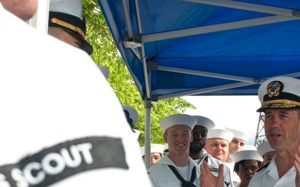 A U.S. Navy officer in dress white uniform with gold-braided cap cover addresses a group of enlisted sailors, also in dress whites, gathered under a blue canopy tent. The foreground shows the back of a sailor's uniform with 'USS SCOUT' printed on the hat band. The setting appears to be an outdoor naval ceremony or briefing event.