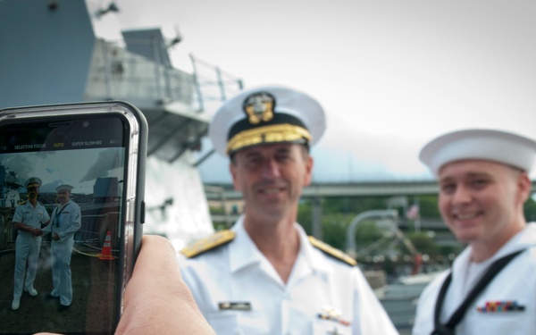 A person is photographing two U.S. Navy personnel using a smartphone on what appears to be the deck of a naval vessel. The officer on the left wears a white service dress uniform with gold-braided cap indicating senior rank, while the sailor on the right wears an enlisted dress white uniform with a Dixie cup hat. The smartphone screen is in sharp focus in the foreground, showing the two subjects being photographed.