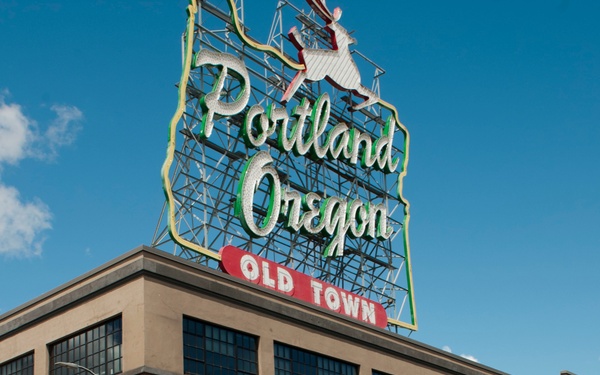 A group of U.S. Navy sailors in white dress uniforms are marching through Old Town Portland, Oregon, likely during a Fleet Week parade or similar military event. The iconic 'Portland Oregon Old Town' neon sign is prominently visible atop a brick building in the background. Several sailors are smiling and giving thumbs-up gestures toward the camera.