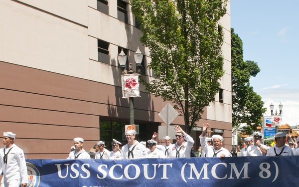 A group of U.S. Navy sailors dressed in white uniforms are marching in a street parade carrying a large blue banner reading 'USS Scout (MCM 8) - Pathfinders, We Lead The Way!' A mascot costume resembling a bald eagle accompanies the group on the left side. The parade appears to take place in an urban setting, likely Portland, Oregon, based on visible signage.