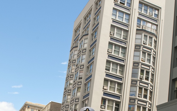 A group of U.S. Navy personnel in white dress uniforms are marching in a street parade carrying a banner for the USS Michael Murphy (DDG-112). The parade takes place on a wet urban street lined with multi-story commercial buildings under a partly cloudy blue sky. A senior enlisted sailor in the foreground is waving to the crowd while an officer marches alongside.