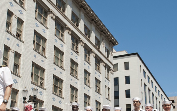 A group of U.S. Navy sailors in dress white uniforms are participating in a public parade, representing the USS Michael Murphy (DDG 112), an Arleigh Burke-class guided-missile destroyer. They are holding a large banner displaying the ship's name, hull number, and motto 'Lead the Fight,' along with a portrait of the ship's namesake. The event takes place on a city street flanked by multi-story buildings under a clear blue sky.