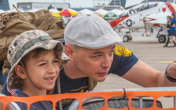 A man and a young child are attending a military air show, standing behind an orange safety barrier fence. The man is pointing at something in the distance, likely directing the child's attention toward aircraft on the tarmac. A U.S. Navy jet aircraft is visible in the background along with other spectators and event tents.