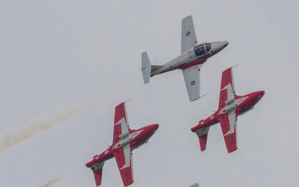 Four military jet aircraft are performing a formation aerobatic display against an overcast grey sky. Two of the aircraft are predominantly red and white, while two appear mostly white with red markings, consistent with the Canadian Forces Snowbirds aerobatic team flying CT-114 Tutor jets. Two of the aircraft are trailing white/grey smoke trails, indicating an active aerobatic performance.
