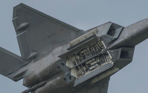 A U.S. Air Force F-22 Raptor stealth fighter jet is photographed from below during flight, with its main weapons bay door open and revealing internal missile/weapon rack hardware. The aircraft is in a banked orientation against an overcast gray sky, providing a clear view of its underside and stealth-optimized airframe geometry. This appears to be an airshow demonstration displaying the internal weapons bay capability.