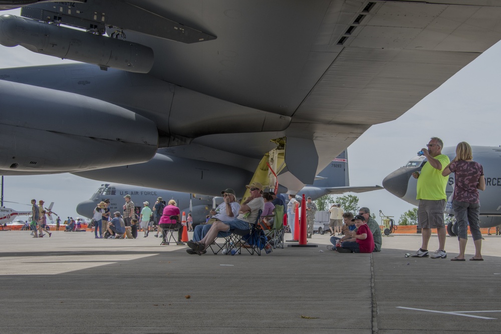 An air show event at a military airfield where civilians are gathered beneath the wings and fuselage of a large U.S. Air Force aircraft, likely a B-52 Stratofortress or similar large bomber/transport. Attendees are relaxing in folding chairs, walking around, and photographing the aircraft on display. Multiple U.S. Air Force aircraft are visible in the background, including what appears to be a C-130 Hercules.