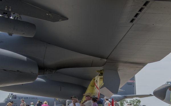 An air show event at a military airfield where civilians are gathered beneath the wings and fuselage of a large U.S. Air Force aircraft, likely a B-52 Stratofortress or similar large bomber/transport. Attendees are relaxing in folding chairs, walking around, and photographing the aircraft on display. Multiple U.S. Air Force aircraft are visible in the background, including what appears to be a C-130 Hercules.