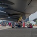 An air show event at a military airfield where civilians are gathered beneath the wings and fuselage of a large U.S. Air Force aircraft, likely a B-52 Stratofortress or similar large bomber/transport. Attendees are relaxing in folding chairs, walking around, and photographing the aircraft on display. Multiple U.S. Air Force aircraft are visible in the background, including what appears to be a C-130 Hercules.