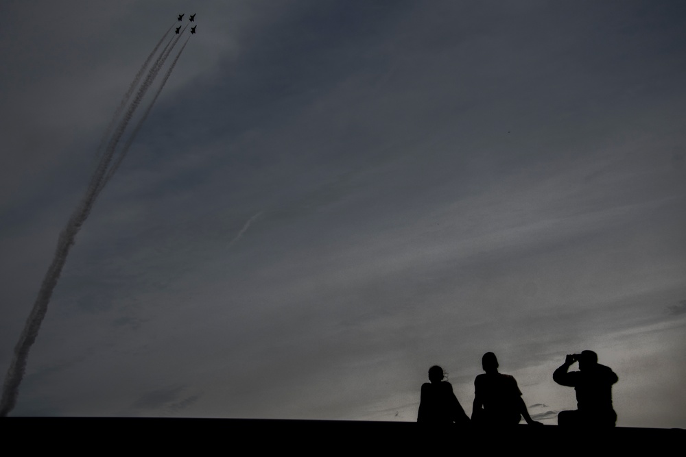 A dramatic silhouette photograph showing three people watching a formation of jet aircraft performing an aerobatic display. The jets, flying in tight formation, leave white smoke trails as they climb steeply into an overcast sky. The composition contrasts the small human figures in the lower right against the vast cloudy sky and ascending aircraft in the upper left.