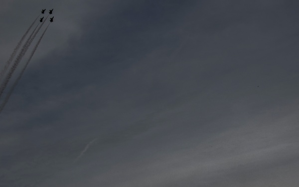 A dramatic silhouette photograph showing three people watching a formation of jet aircraft performing an aerobatic display. The jets, flying in tight formation, leave white smoke trails as they climb steeply into an overcast sky. The composition contrasts the small human figures in the lower right against the vast cloudy sky and ascending aircraft in the upper left.