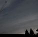 A dramatic silhouette photograph showing three people watching a formation of jet aircraft performing an aerobatic display. The jets, flying in tight formation, leave white smoke trails as they climb steeply into an overcast sky. The composition contrasts the small human figures in the lower right against the vast cloudy sky and ascending aircraft in the upper left.