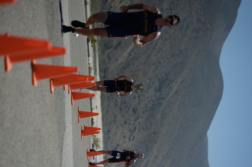 The image is rotated 90 degrees, showing athletes running along a road lined with orange traffic cones. The runners appear to be participating in a triathlon or road race, wearing matching dark athletic uniforms. A mountainous desert landscape is visible in the background under a clear sky.