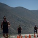 The image is rotated 90 degrees, showing athletes running along a road lined with orange traffic cones. The runners appear to be participating in a triathlon or road race, wearing matching dark athletic uniforms. A mountainous desert landscape is visible in the background under a clear sky.