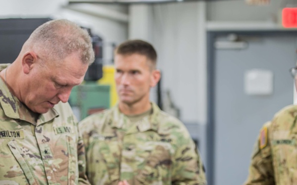 A group of U.S. Army soldiers in OCP (Operational Camouflage Pattern) uniforms are gathered in what appears to be a maintenance or industrial facility. The central figure, identified by nametape as 'HAMILTON,' is examining a small mechanical component, likely a gear or sprocket. The scene appears to be an inspection or briefing visit.