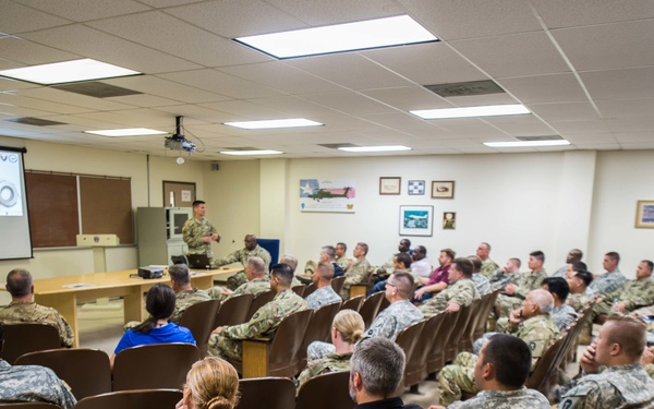 A military briefing or training session is taking place in a classroom-style auditorium with tiered seating. The majority of attendees are wearing U.S. Army combat uniforms (ACU pattern), with a presenter standing at the front near a projected presentation screen. A small number of civilians are also present among the audience.