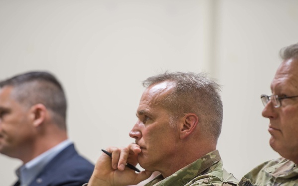 The image shows a group of individuals seated in what appears to be a briefing or meeting room. Two individuals in the foreground are wearing military camouflage uniforms (OCP/MultiCam pattern), while a man in a dark business suit is visible in the background. The subjects appear to be attentively listening to a presentation or speaker off-camera.