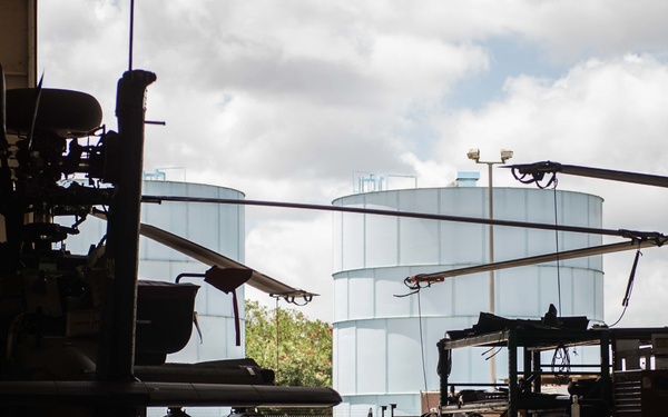 The image shows the interior of a military aviation maintenance hangar with at least two military helicopters visible in silhouette. The hangar opens to an outdoor area where large cylindrical storage tanks (likely fuel tanks) are visible in the background. The scene suggests active maintenance operations with personnel and equipment present.
