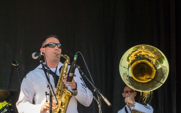 Two musicians dressed in white naval uniforms are performing on an outdoor stage. The foreground musician plays an alto saxophone while the background musician plays a sousaphone (tuba). A drum kit and stage equipment are visible in the background.