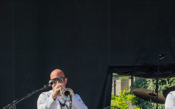 A bald man wearing a white suit and sunglasses is playing a trumpet on an outdoor stage during a live performance. He is positioned in front of a microphone stand, with stage equipment, amplifiers, and a large electric fan visible in the background. A second performer in white is partially visible on the right edge of the frame.