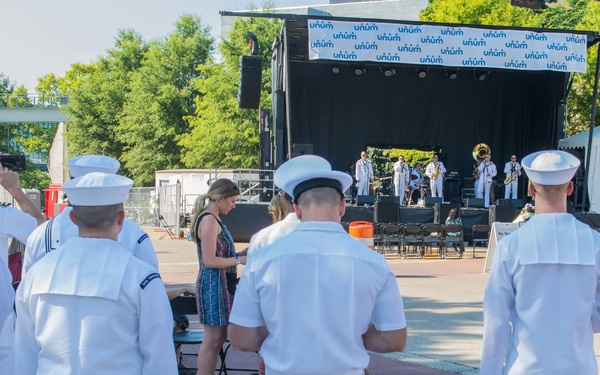A group of U.S. Navy sailors in white dress uniforms are watching an outdoor concert performance on a stage. The stage backdrop displays repeated 'Unum' branding, and a band with brass instruments is performing. The event appears to be a public outdoor venue on a sunny day.