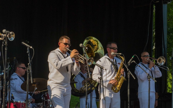 A military band, likely U.S. Navy based on the distinctive white dress uniforms, performs on an outdoor stage. The ensemble includes brass and woodwind instruments along with a drum kit, suggesting a full concert band or jazz ensemble performance. The setting appears to be a public outdoor event with stage monitors, microphones, and green trees visible in the background.