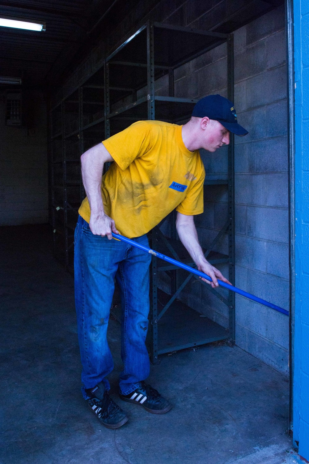 A young man in a yellow t-shirt and blue jeans is working inside what appears to be an empty warehouse or storage facility. He is bent over, gripping a blue-handled tool — likely a broom or mop — and appears to be cleaning or sweeping the concrete floor. The space features empty metal shelving units and cinder block walls.
