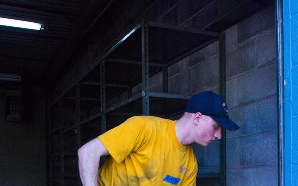 A young man in a yellow t-shirt and blue jeans is working inside what appears to be an empty warehouse or storage facility. He is bent over, gripping a blue-handled tool — likely a broom or mop — and appears to be cleaning or sweeping the concrete floor. The space features empty metal shelving units and cinder block walls.