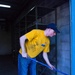 A young man in a yellow t-shirt and blue jeans is working inside what appears to be an empty warehouse or storage facility. He is bent over, gripping a blue-handled tool — likely a broom or mop — and appears to be cleaning or sweeping the concrete floor. The space features empty metal shelving units and cinder block walls.