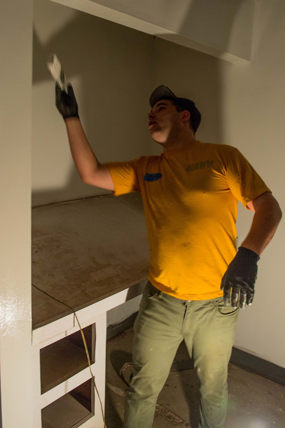 A young man wearing a yellow t-shirt, green cargo pants, and black work gloves is performing construction or renovation work inside a room. He is reaching upward with his left hand, appearing to apply material or inspect a wall or ceiling surface. The setting shows an unfinished or partially renovated interior space with white-painted structures and exposed flooring.