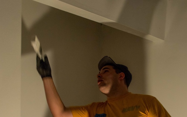 A young man wearing a yellow t-shirt, green cargo pants, and black work gloves is performing construction or renovation work inside a room. He is reaching upward with his left hand, appearing to apply material or inspect a wall or ceiling surface. The setting shows an unfinished or partially renovated interior space with white-painted structures and exposed flooring.