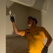 A young man wearing a yellow t-shirt, green cargo pants, and black work gloves is performing construction or renovation work inside a room. He is reaching upward with his left hand, appearing to apply material or inspect a wall or ceiling surface. The setting shows an unfinished or partially renovated interior space with white-painted structures and exposed flooring.